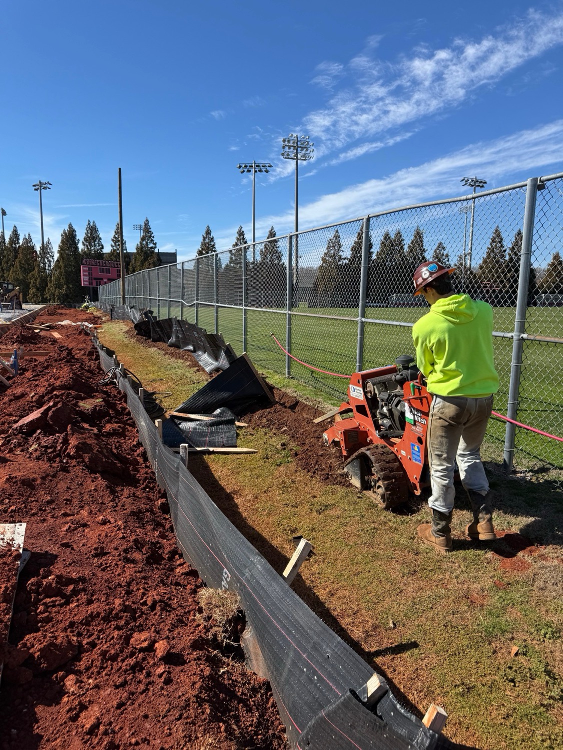 Crew member trenching in a silt fence line with a mini skid steer along a sports complex